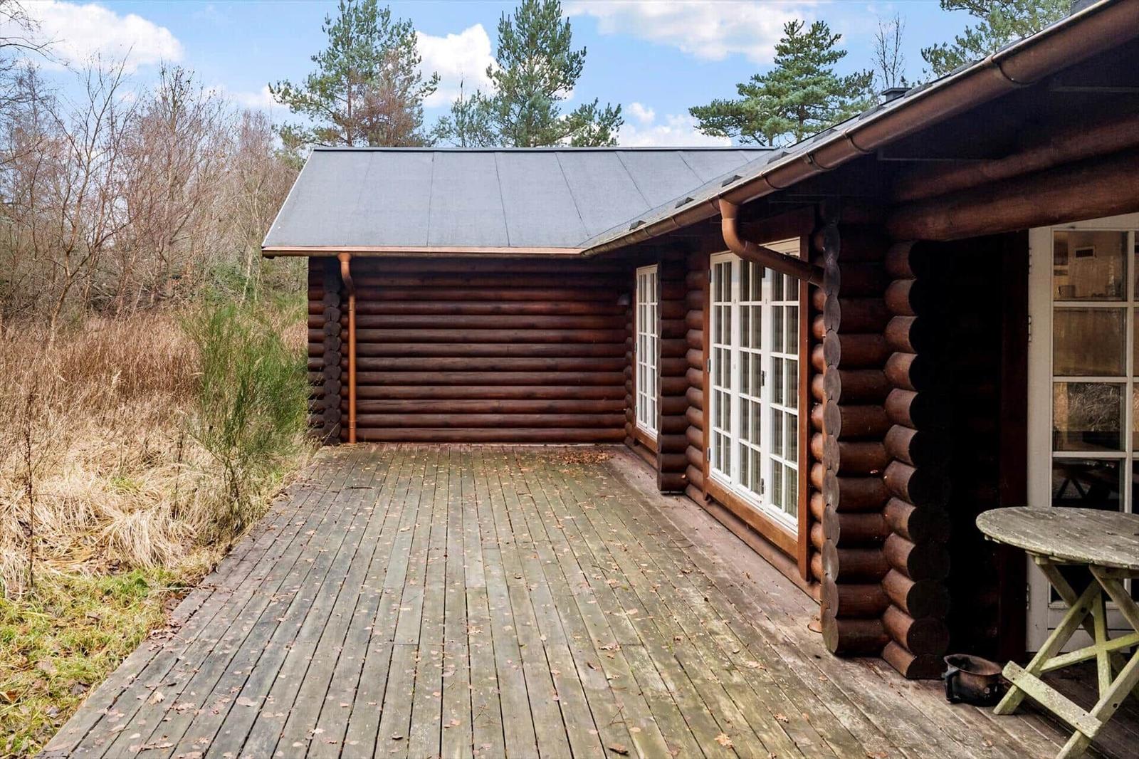 Holzhaus mit Terrasse und Fenstern. Umgeben von Bäumen und Gras.