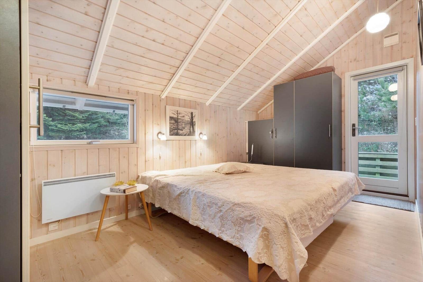 Bedroom with wooden walls, bed, wardrobe, and window with view of forest.