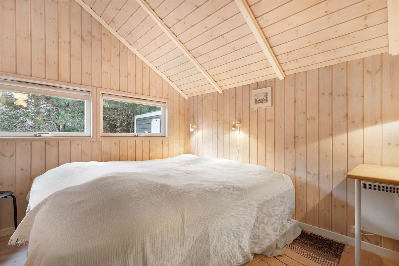 Bedroom with wooden walls and sloped ceiling, bed, windows, and desk.