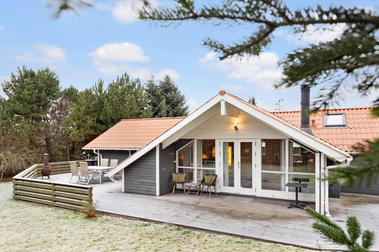 Modern wooden cabin with terrace, seating, and grill area in green surroundings.