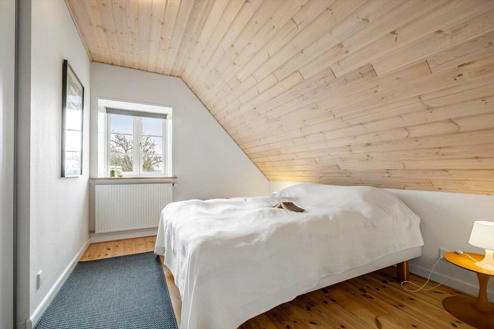 Schlafzimmer mit Holzdecke, weißem Bett und Fenster mit Blick nach draußen.