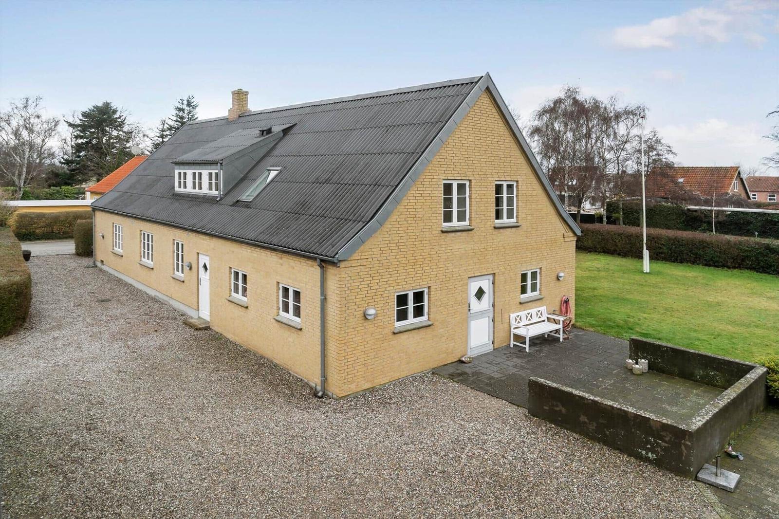 Ein gelbes Haus mit grauem Dach und weißem Fensterrahmen. Vor dem Haus befindet sich eine Terrasse mit weißem Stuhl.