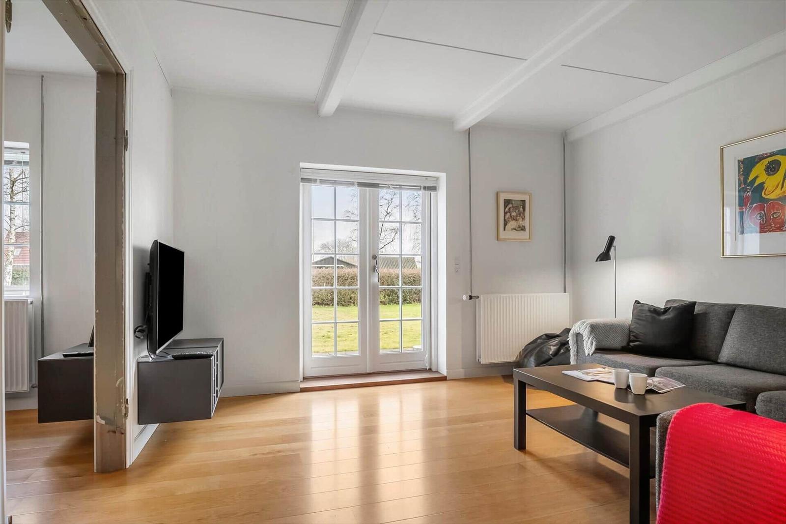 Wohnzimmer mit Sofa, Couchtisch und TV. Durchgang zur Küche. Fenster zur grünen Außenseite.