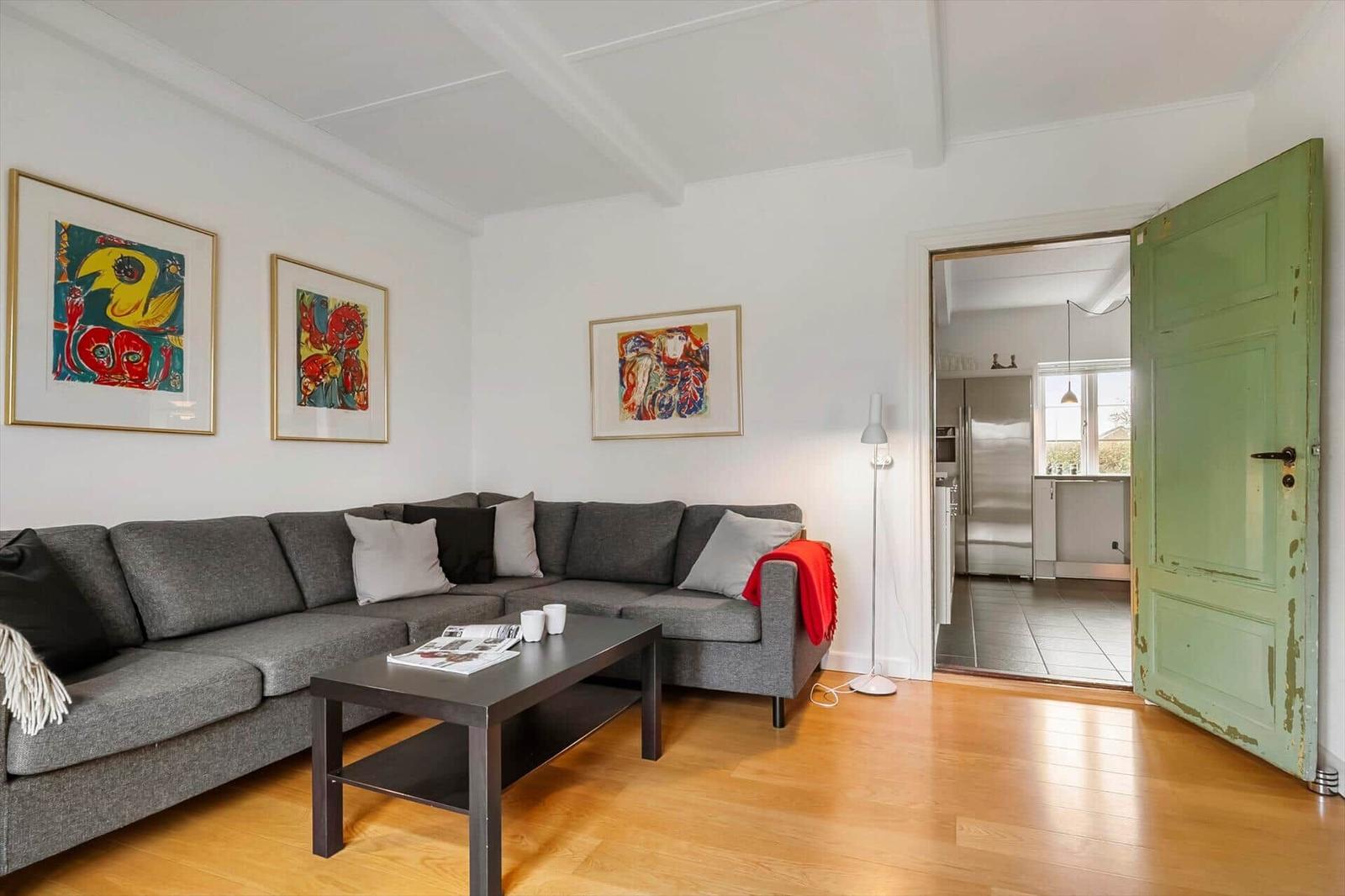 Wohnzimmer mit grauem Sofa, Holzboden und drei Wandbildern. Grüne Tür führt in die Küche.