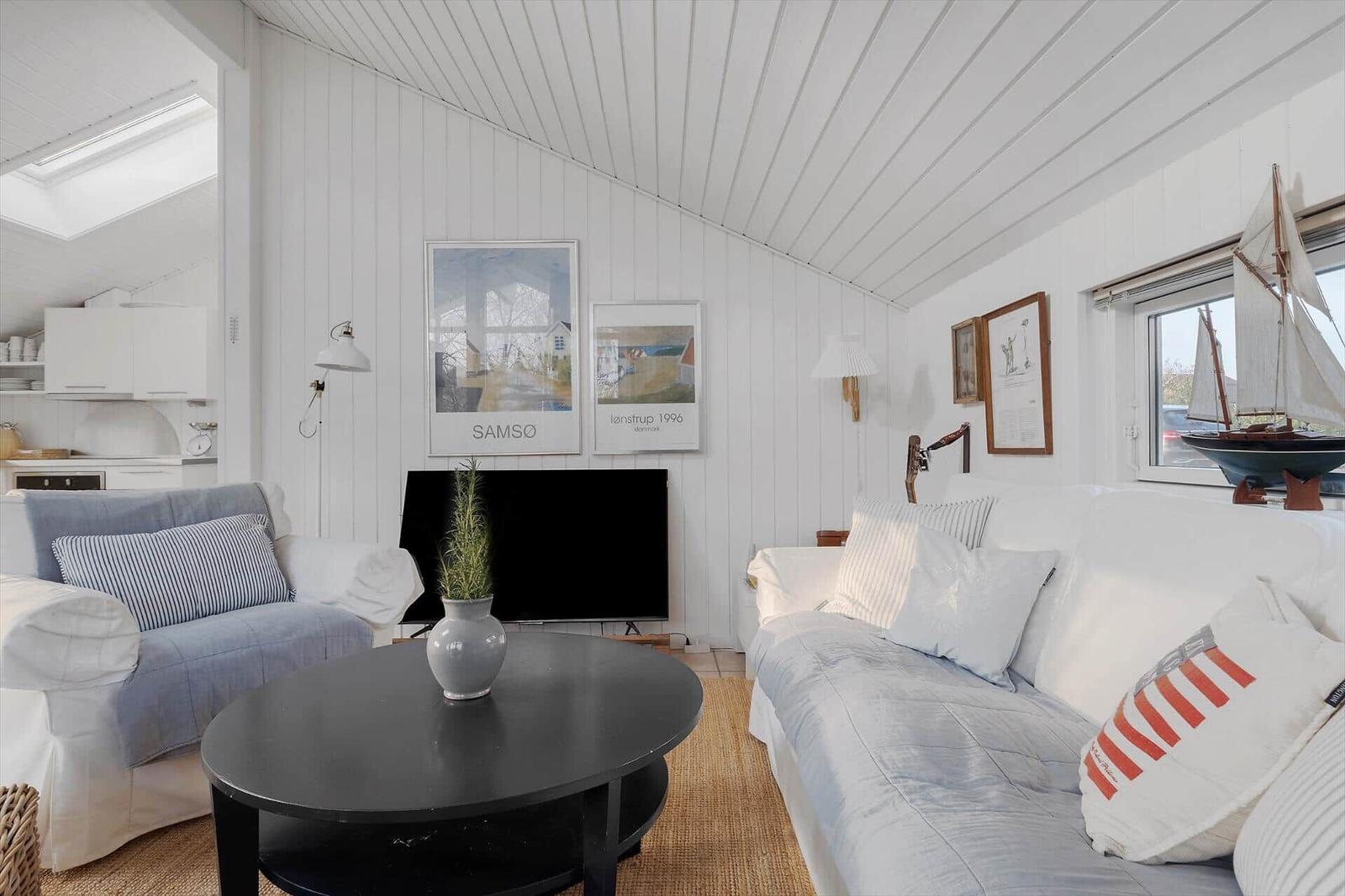 Living room with sofa, armchair, and round table. Posters hang on the wall, and a sailboat model stands by the window.