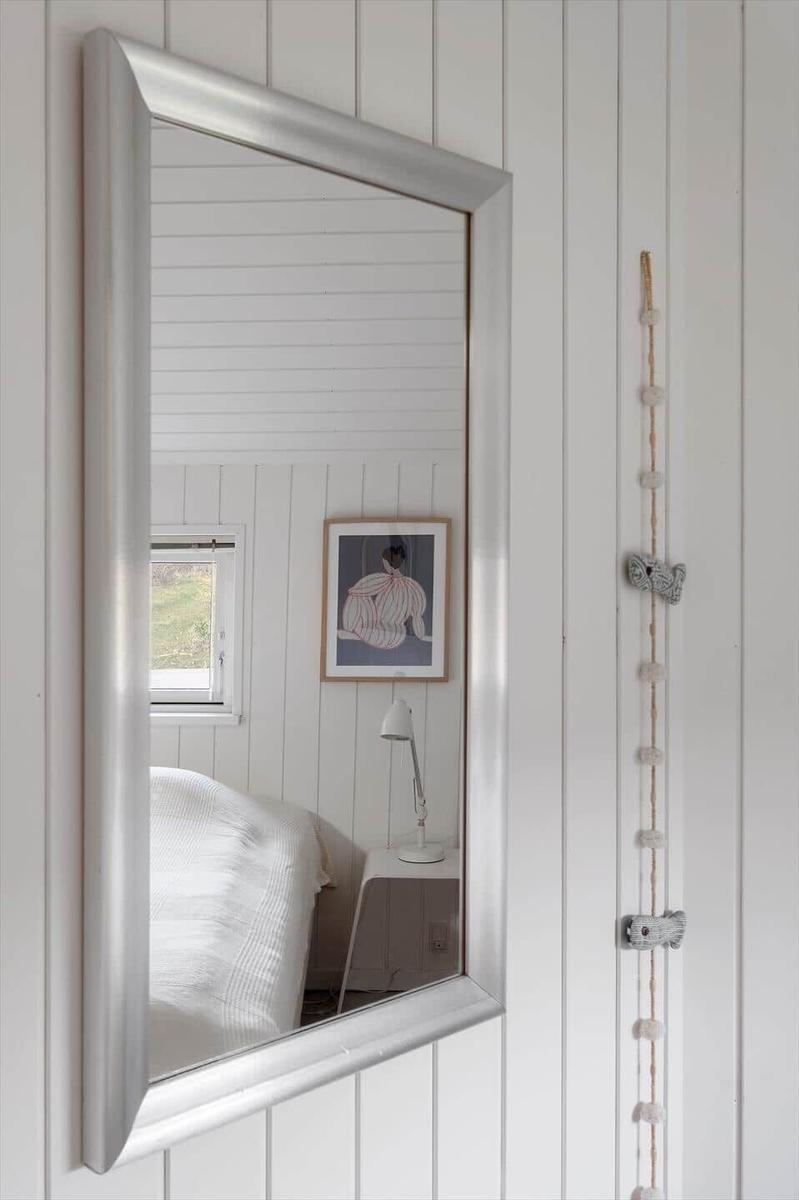 Mirror reflects bed, window, and wall art in white-paneled room.