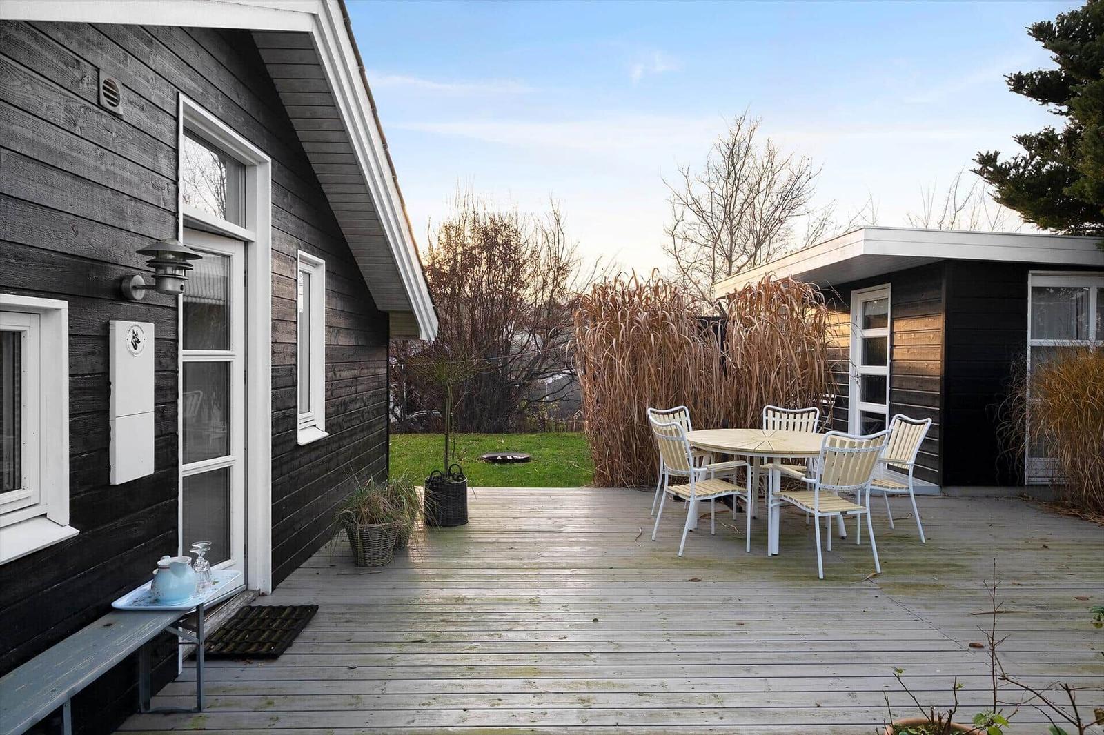 Deck with table and chairs in front of black wooden house with garden.