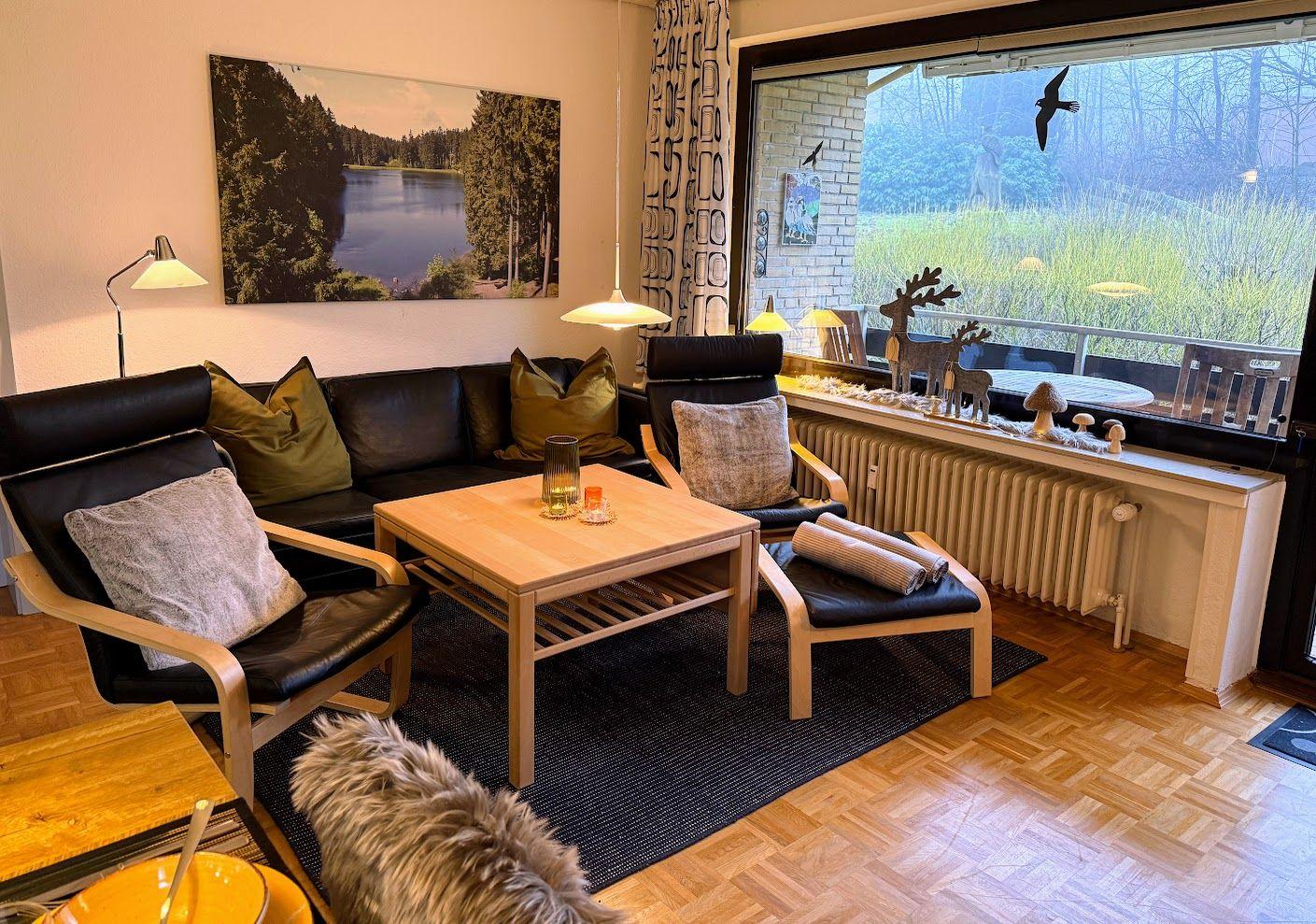 Wohnzimmer mit Sofa, Sessel und Tisch. Große Fensterfront mit Blick auf Grünfläche.