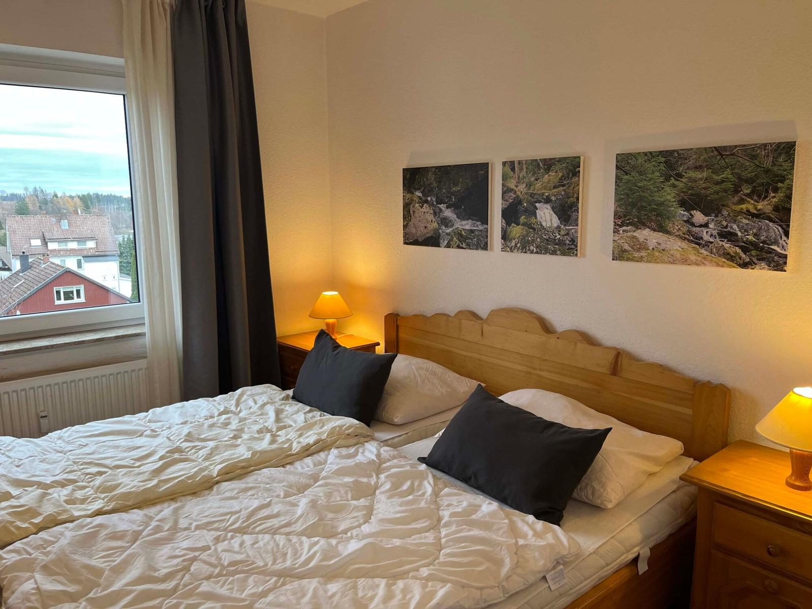 Schlafzimmer mit Bett, Nachttischen und Fensterblick auf Dächer.