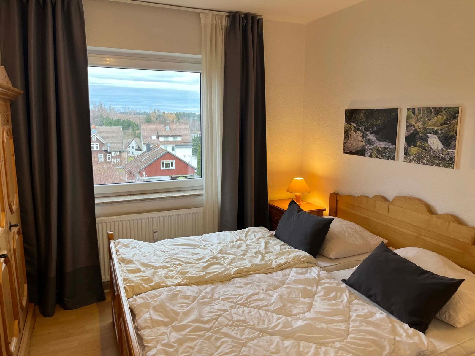 Schlafzimmer mit Blick auf Dachdörfer und Wald. Holzbett, Nachttisch mit Lampe und Bilder an der Wand.