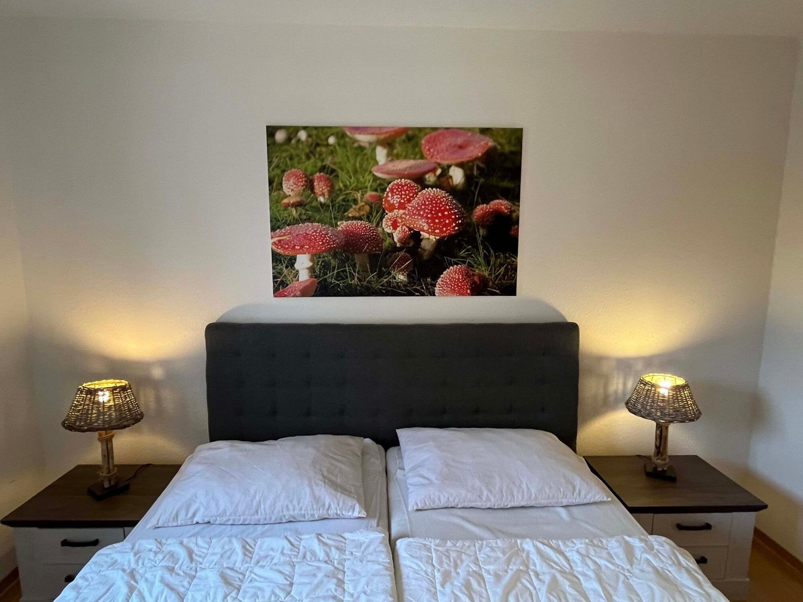 Schlafzimmer mit Bett, Nachttischen und Wandbild mit Pilzen.