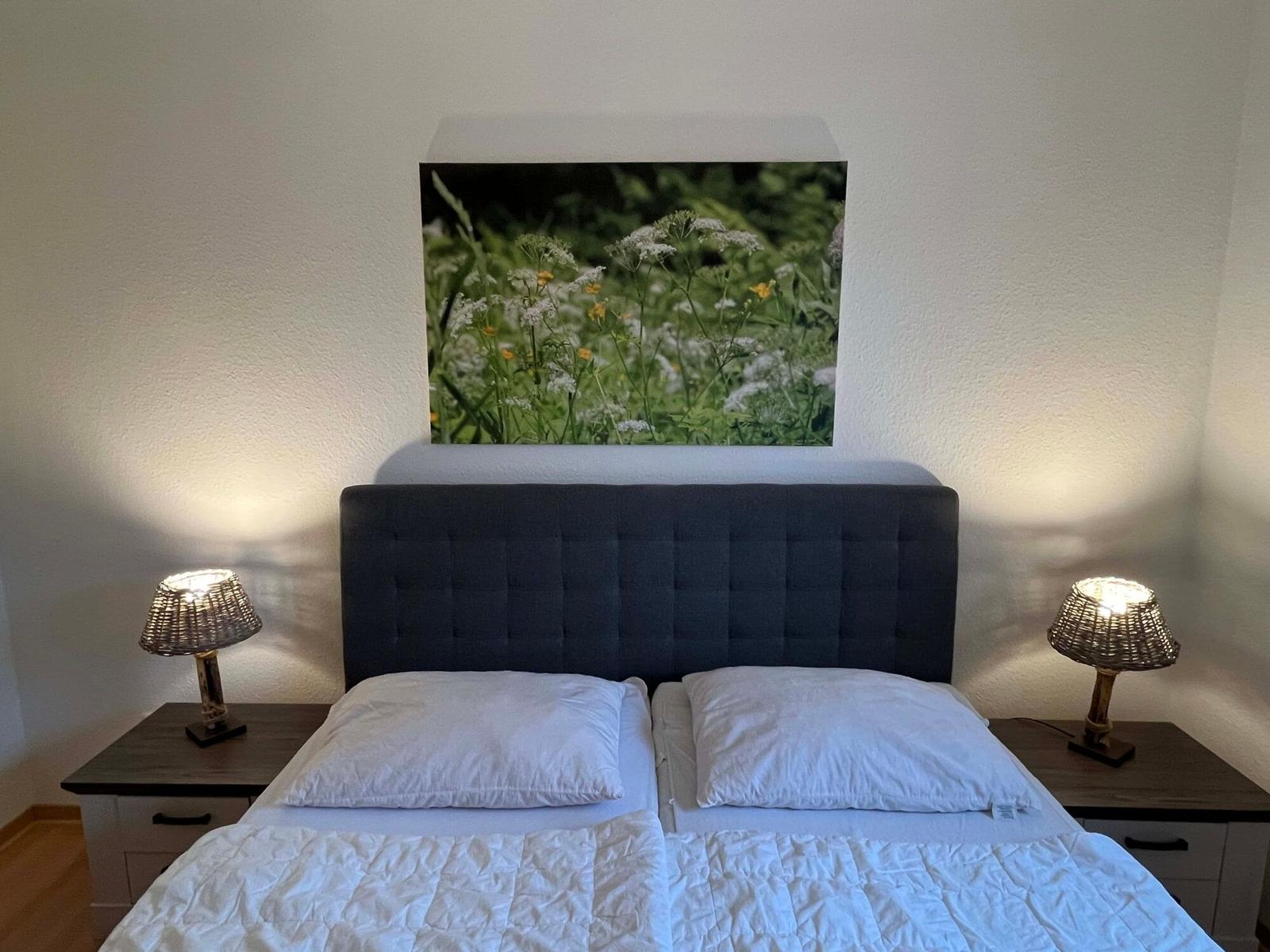 Schlafzimmer mit Doppelbett, zwei Nachttischen und Wandbild mit Blumen.