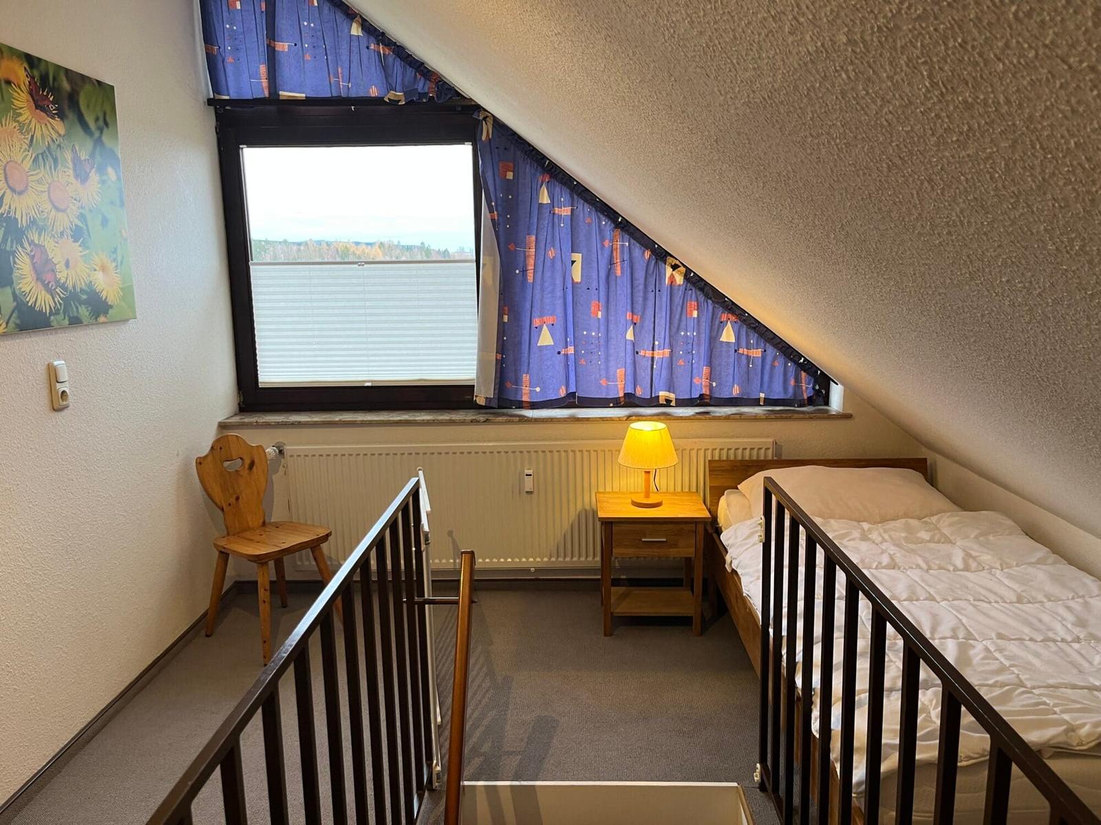 Schlafzimmer mit Dachschräge, Bett, Nachttisch und Fenster mit Vorhängen.