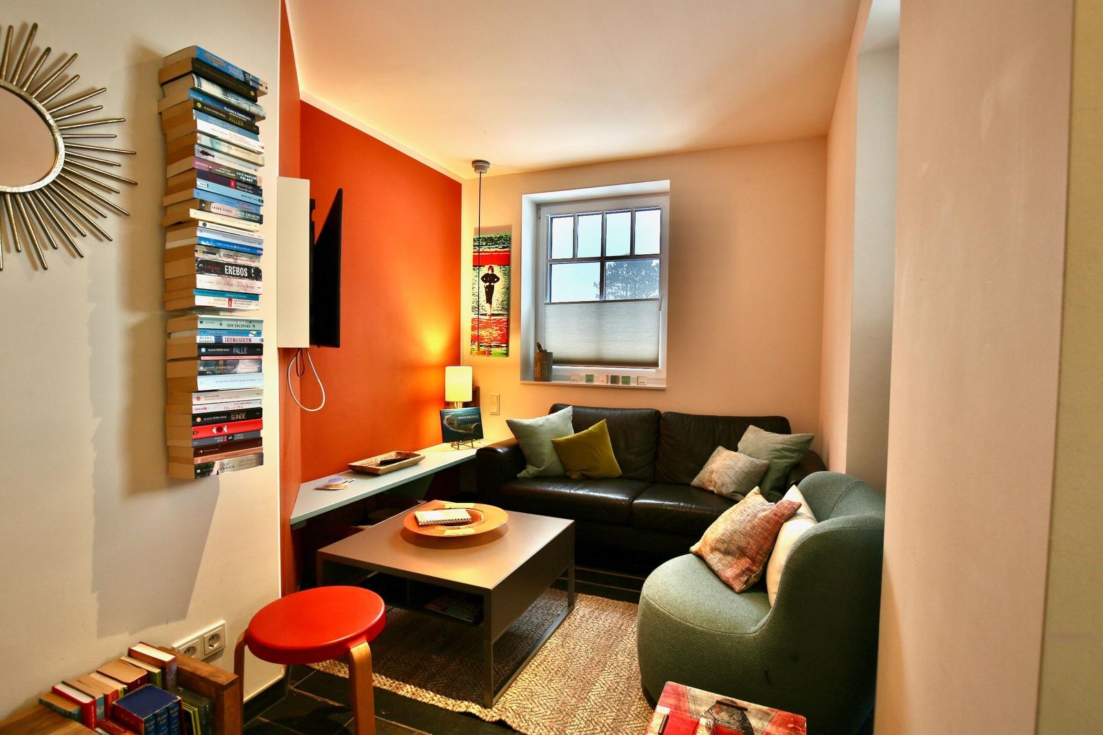 Wohnzimmer mit Sofa, Sessel und Couchtisch. Wandregal mit Büchern und Fenster.