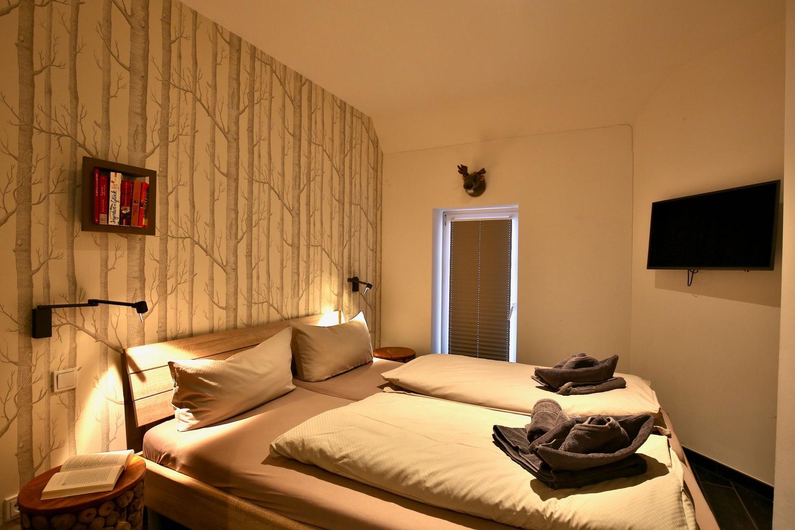 Zimmer mit Doppelbett, Waldtapete, Fernseher und Wandregal mit Büchern.