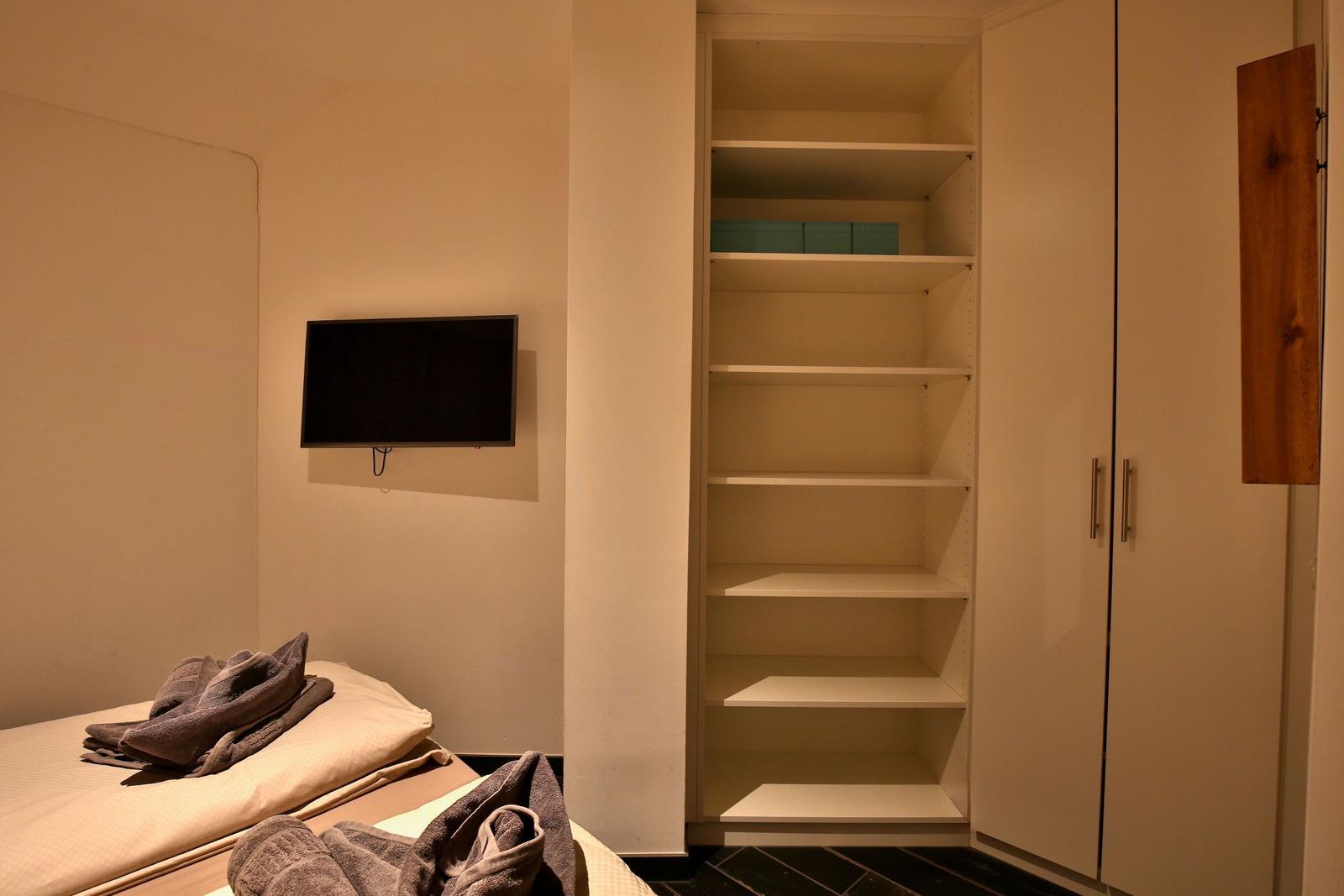 Schlafzimmer mit Bett, TV und Kleiderschrank mit Regalen.