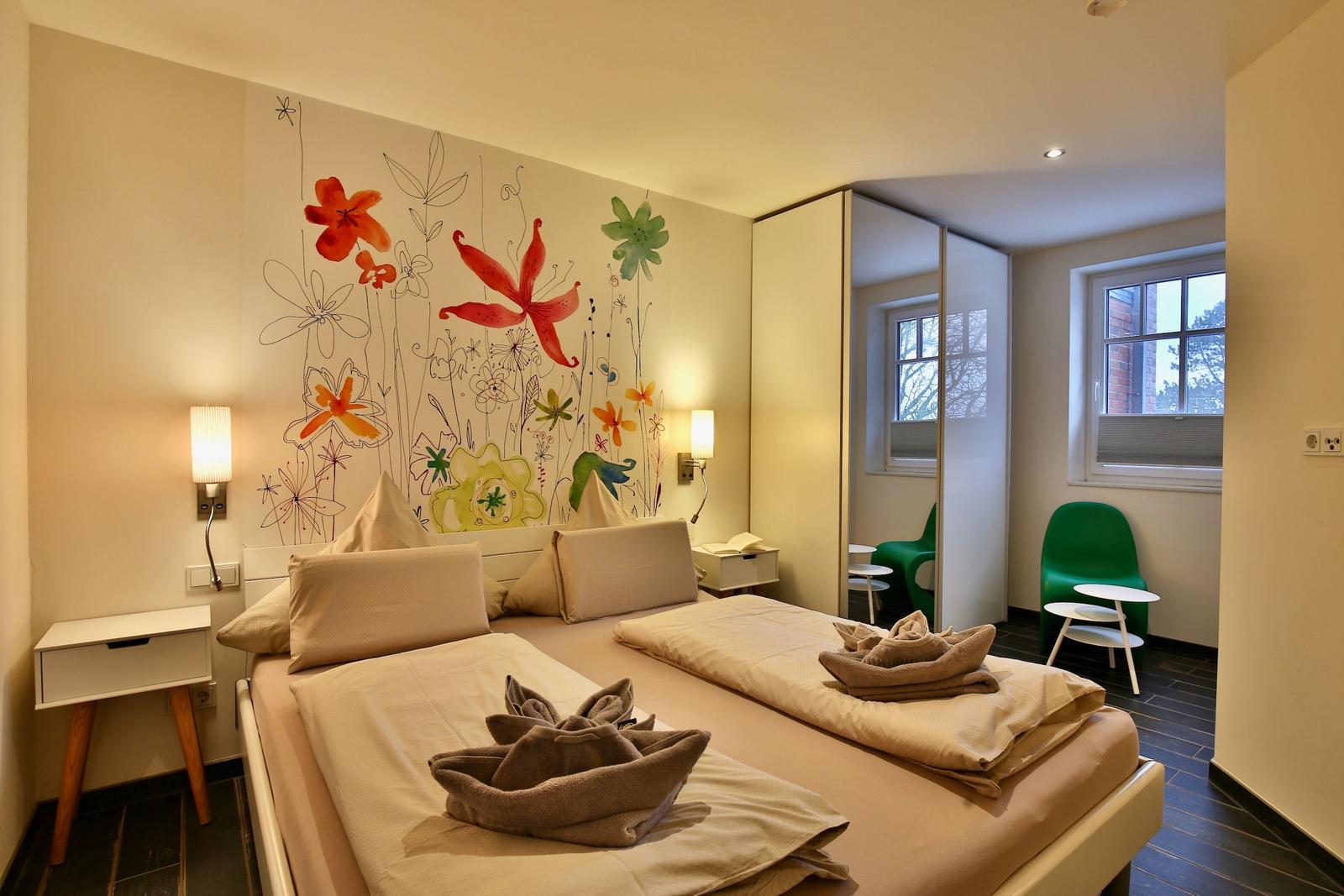 Schlafzimmer mit Bett, Wandmalerei und grüner Sitzgelegenheit.