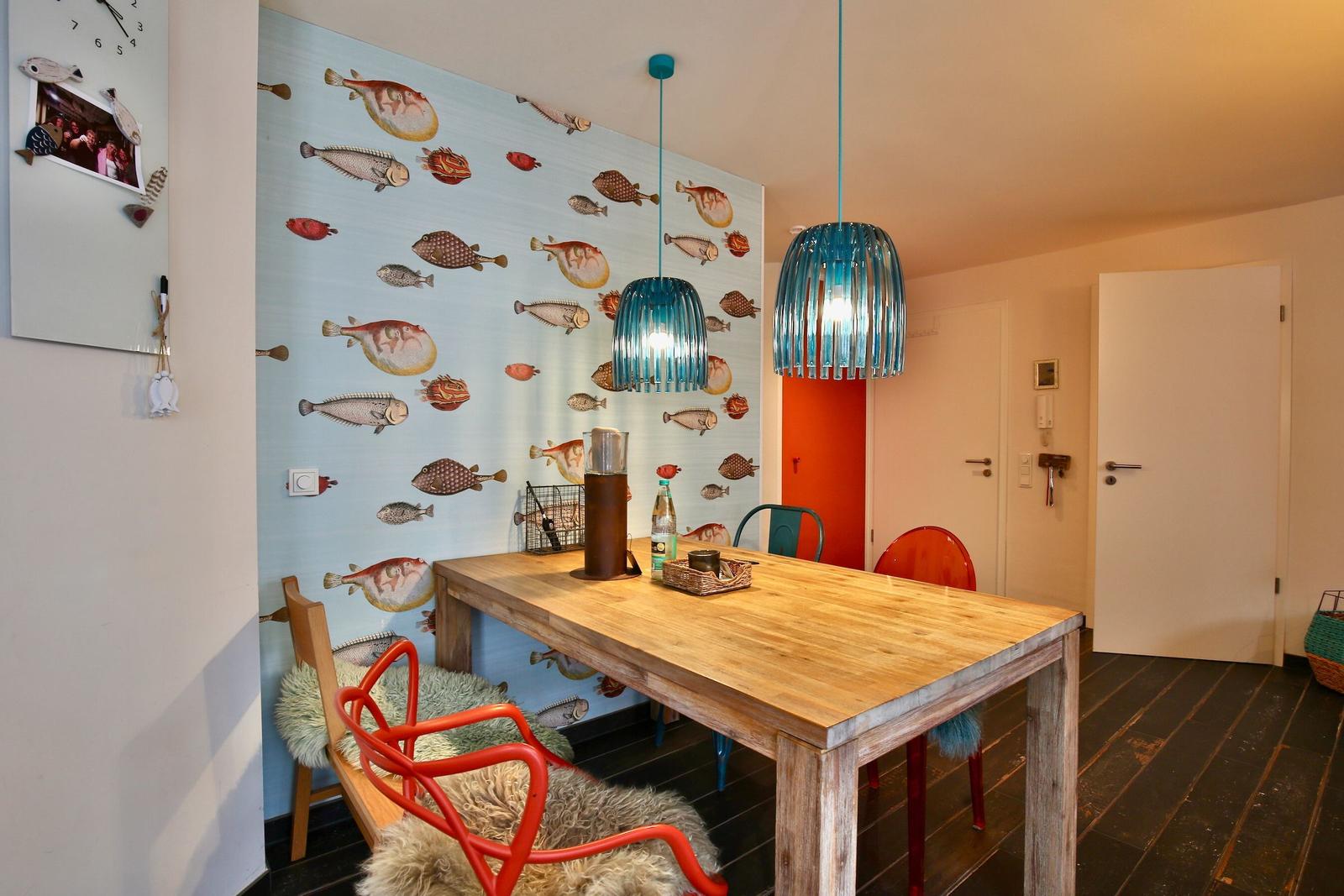 Essbereich mit Holztisch, bunten Stühlen und Fischmotiv-Wandtapete.