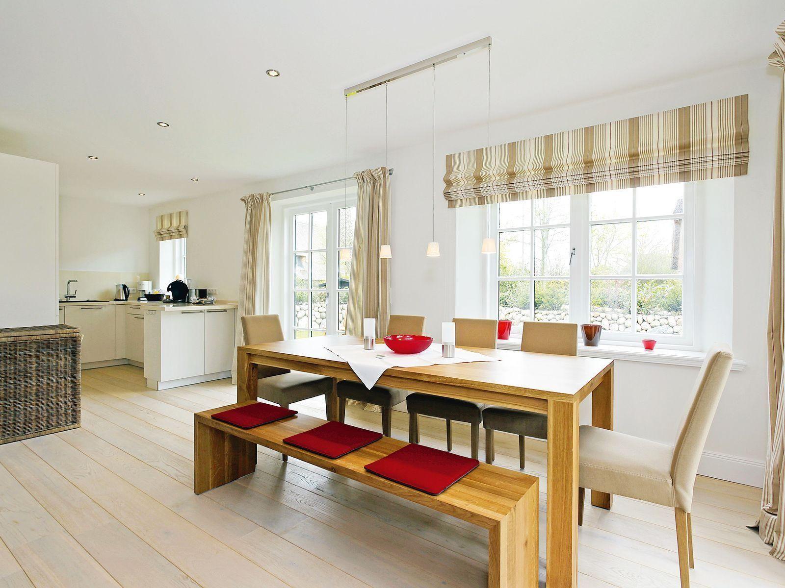 Dining area with wooden table, benches, and chairs. Kitchen in background. Windows with blinds.