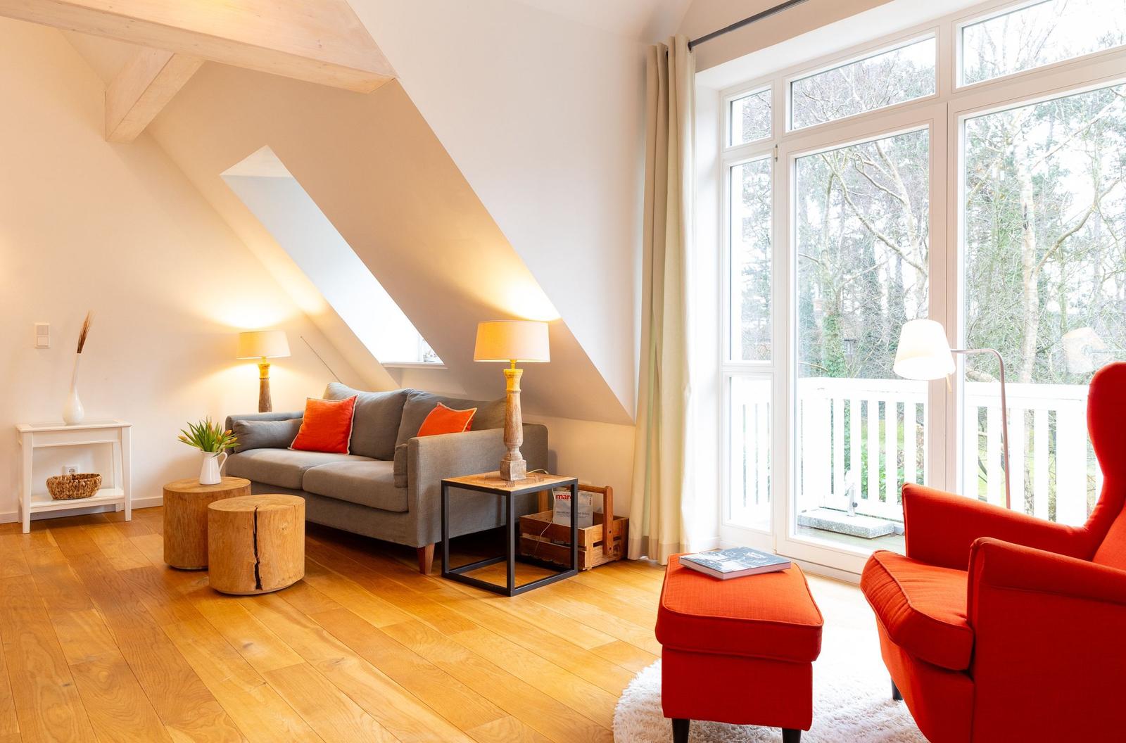 Wohnzimmer mit grauem Sofa, rotem Sessel und Holzboden. Große Fenster mit Blick auf den Garten.