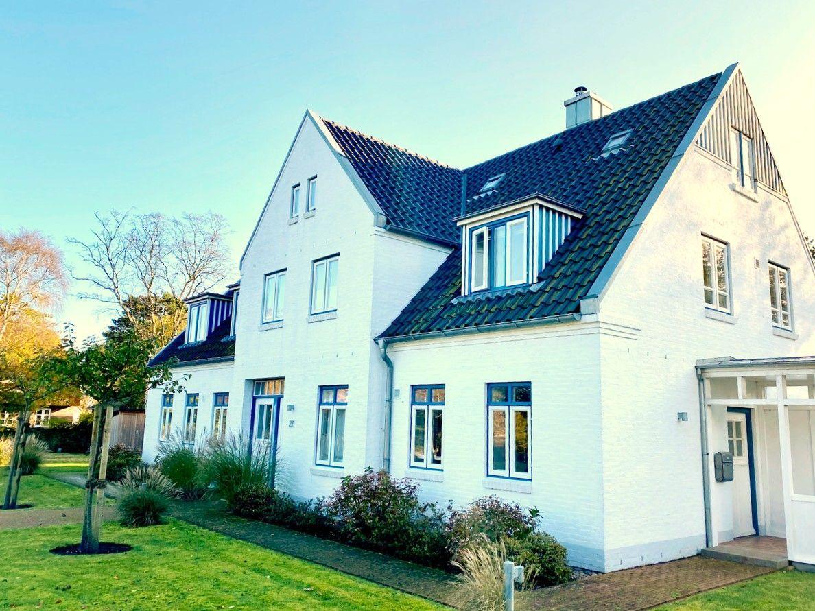 Weiße Villa mit blauen Fensterrahmen und dunklem Dach, umgeben von Grünfläche und Bäumen.