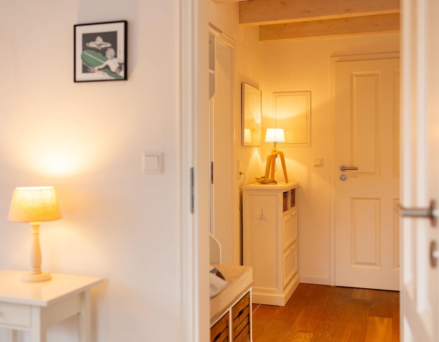 Flur mit Holzboden, weißen Wänden und einer Tür. Eine Lampe steht auf einem Schrank.