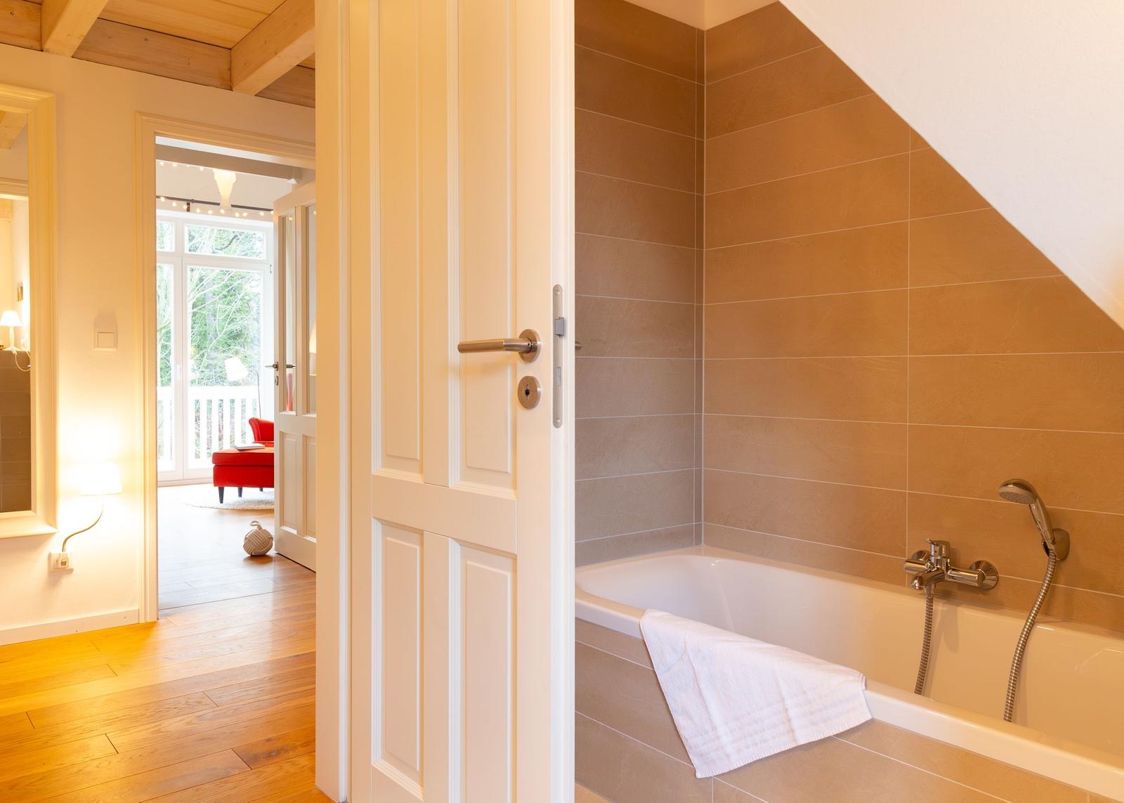 Bad mit Badewanne und Duschbrause. Tür öffnet sich in Wohnbereich mit Holzboden.