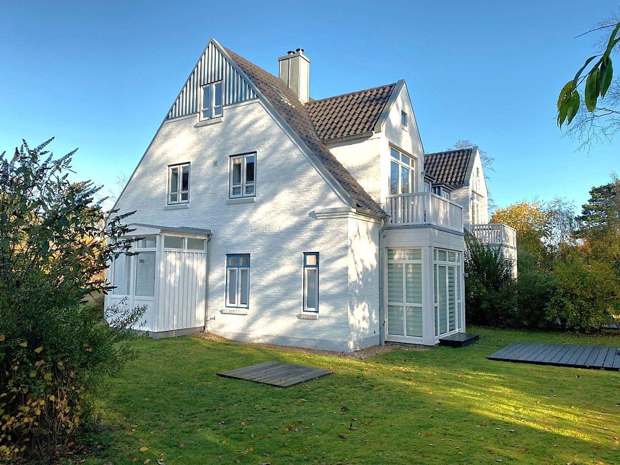 Weißes Haus mit Balkon und grünem Garten unter blauem Himmel.