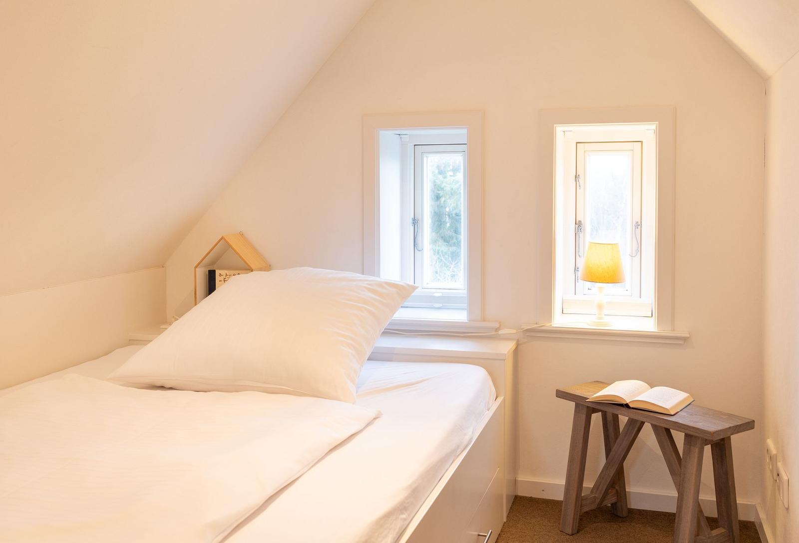 Schlafzimmer mit Bett, zwei Fenstern und Holztisch mit Buch.