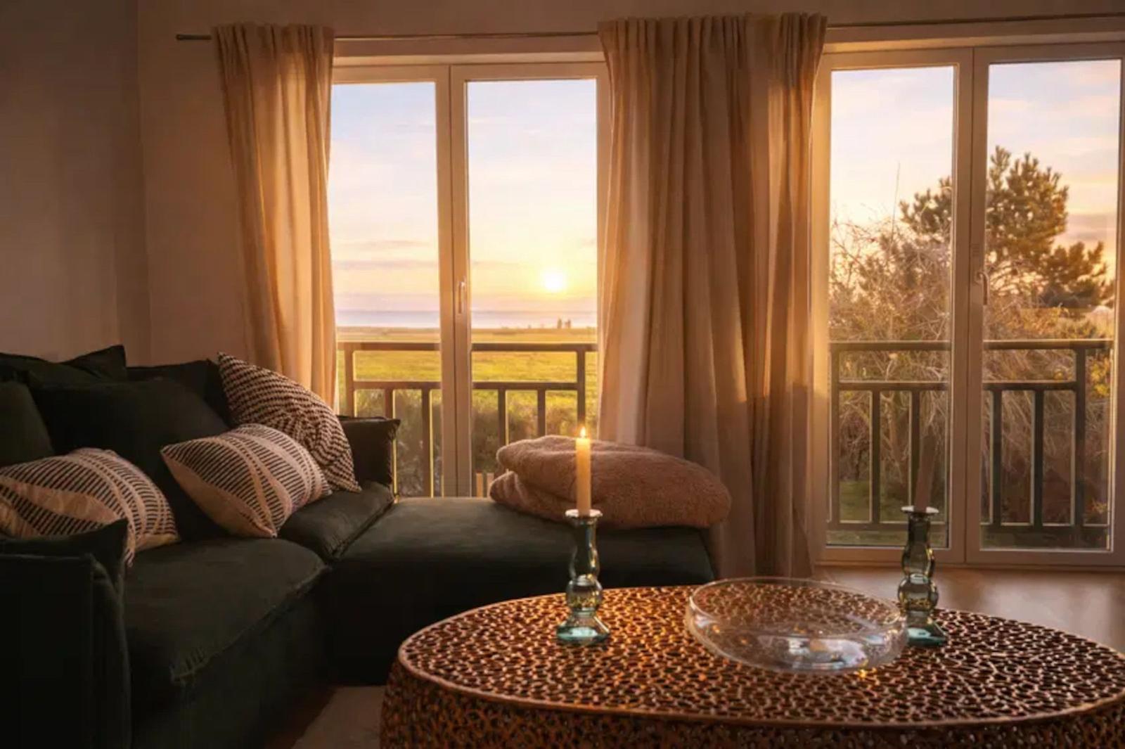 Wohnzimmer mit Sofa und Tisch. Durch Fenster ist Sonnenuntergang über Meer sichtbar.