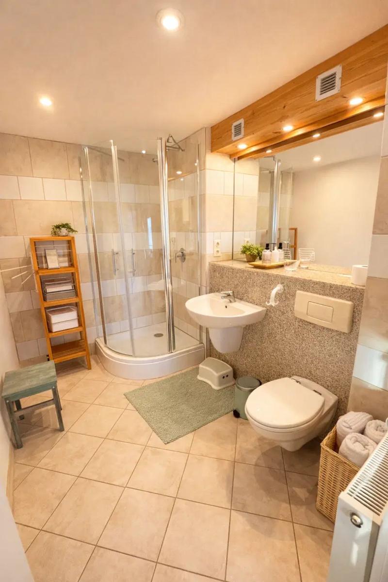 Badezimmer mit Dusche, Waschbecken und Toilette. Holzregal und Handtuchkorb.