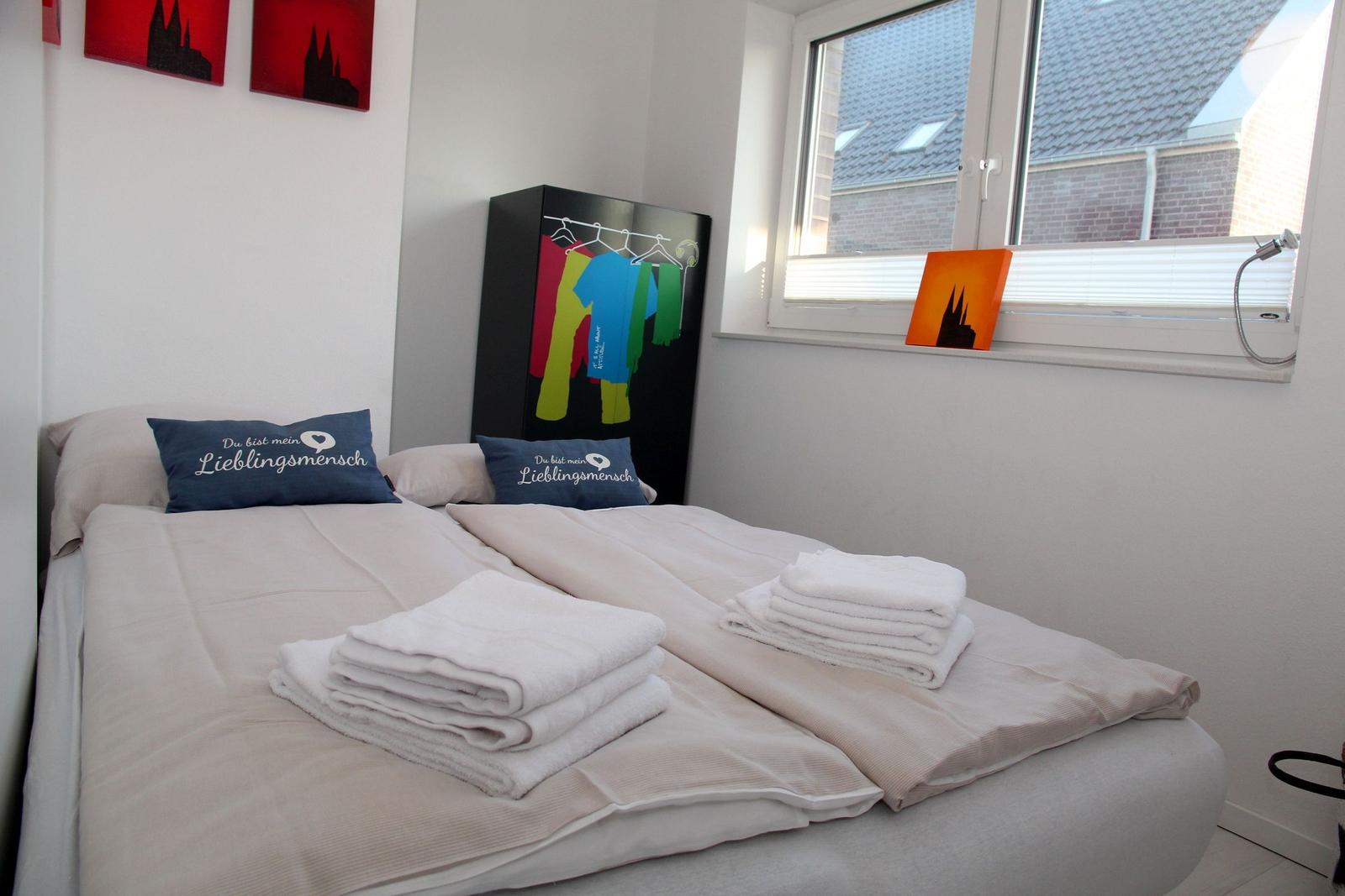 Schlafzimmer mit Bett, Handtüchern und Kleiderschrank. Fenster mit Blick auf Dächer.