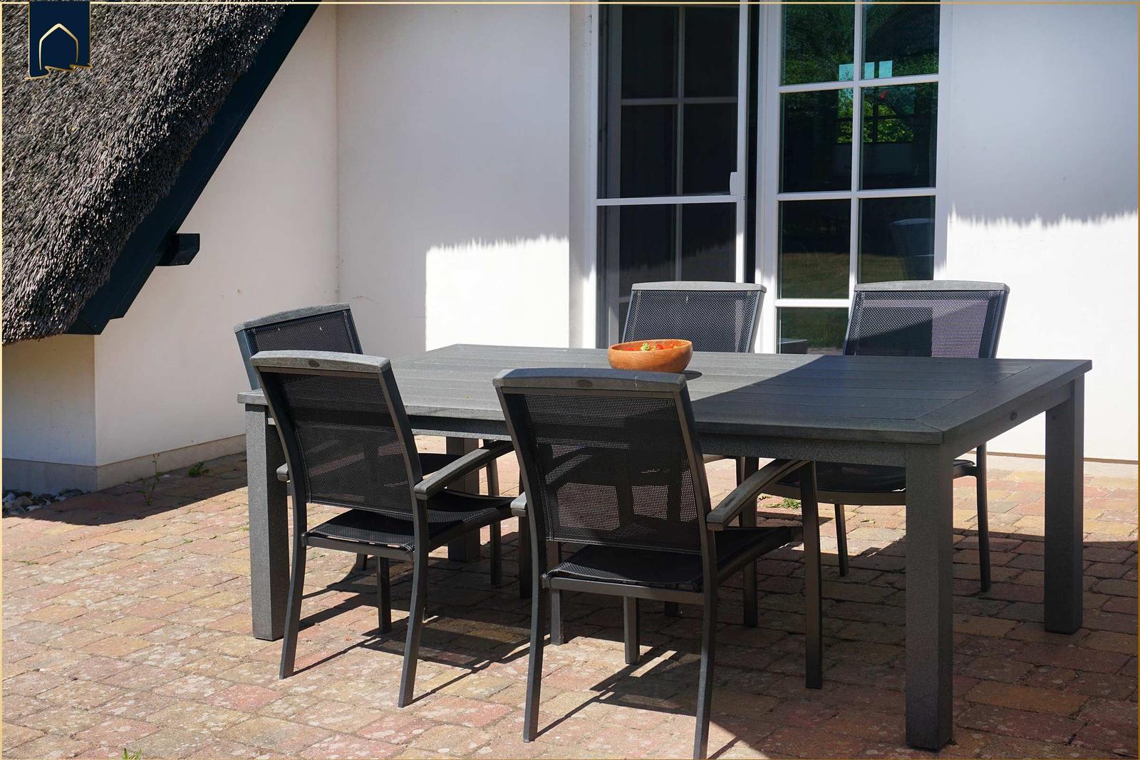 Terrasse mit Tisch und vier Stühlen. Hintergrund: Haus mit Dach und Fenster.