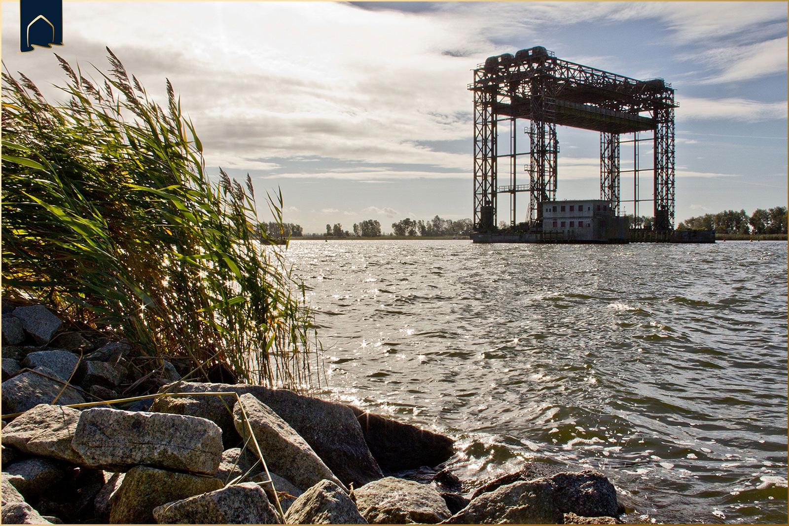 Wasserufer mit Steinen und Schilf, im Hintergrund ein Hafenkran.