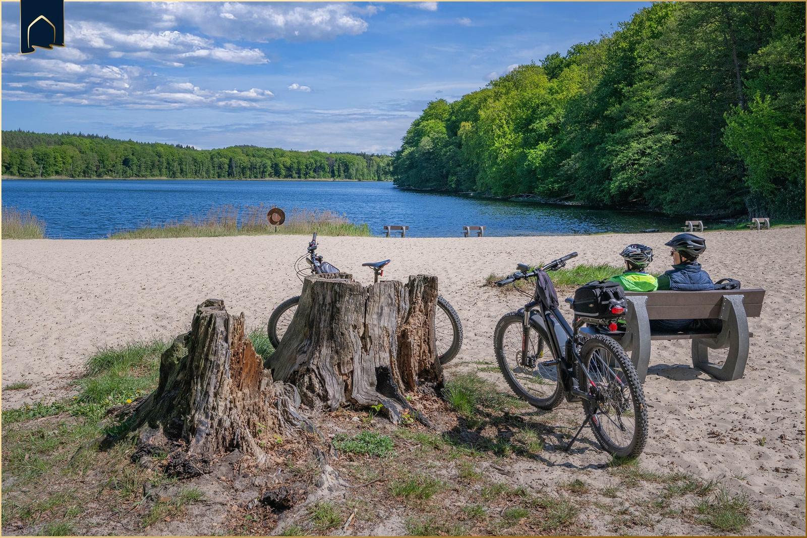 Zwei Radfahrer sitzen auf einer Bank am Strand. Ein Fahrrad steht daneben. Im Hintergrund liegt ein See.
