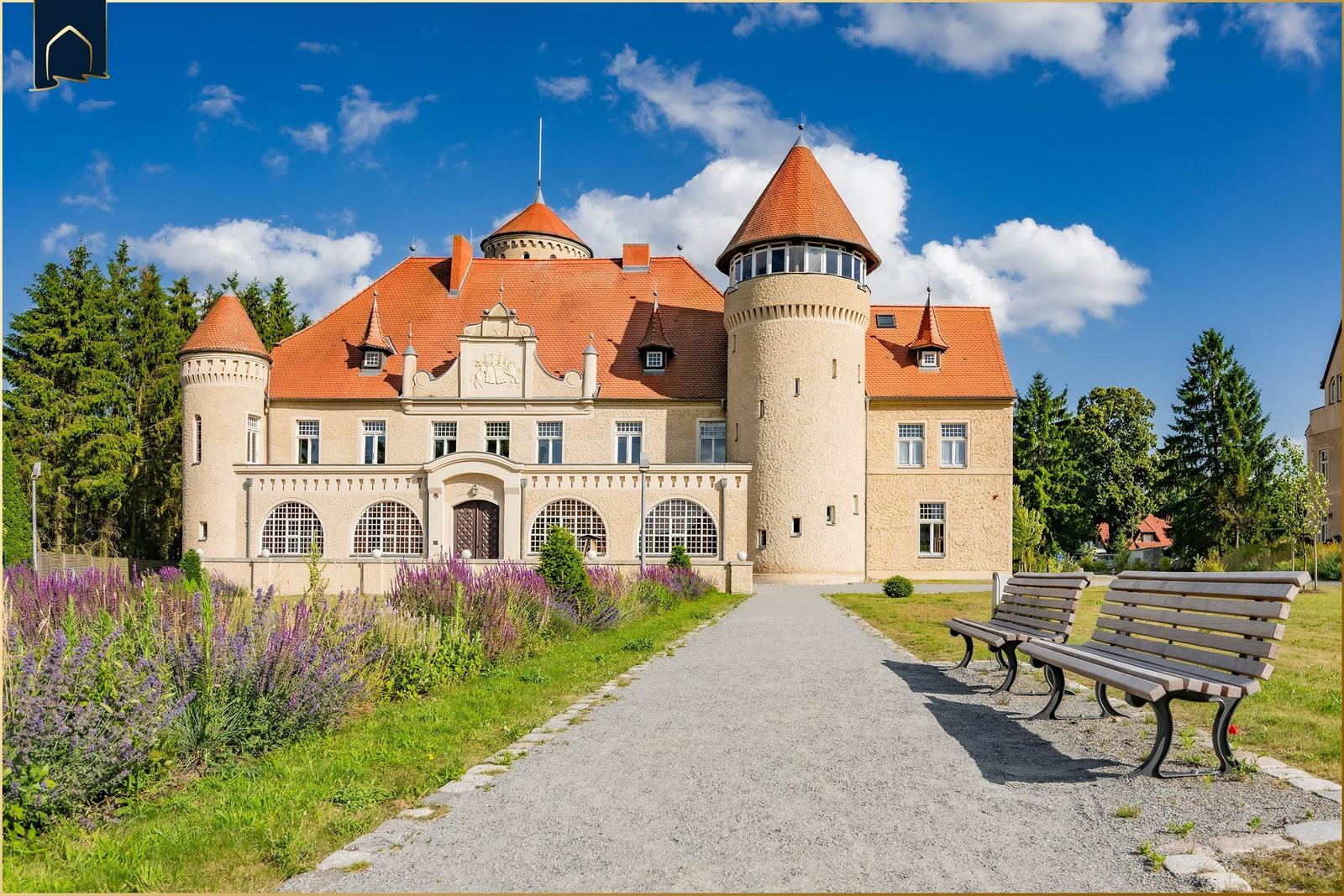 Schloss mit rotem Dach und zwei Türmen. Vorhof mit Bank und Blumenbeete.