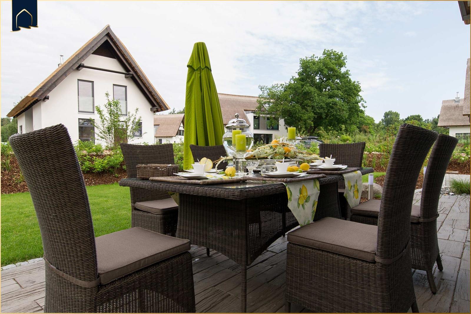 Garten-Terrasse mit gedecktem Esstisch und grünem Sonnenschirm. Hintergrund: weißes Haus mit Dach.