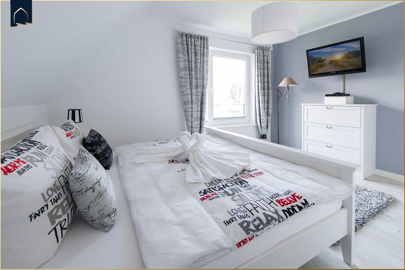 Schlafzimmer mit weißem Bett, Fernseher und Fenster mit Vorhängen.