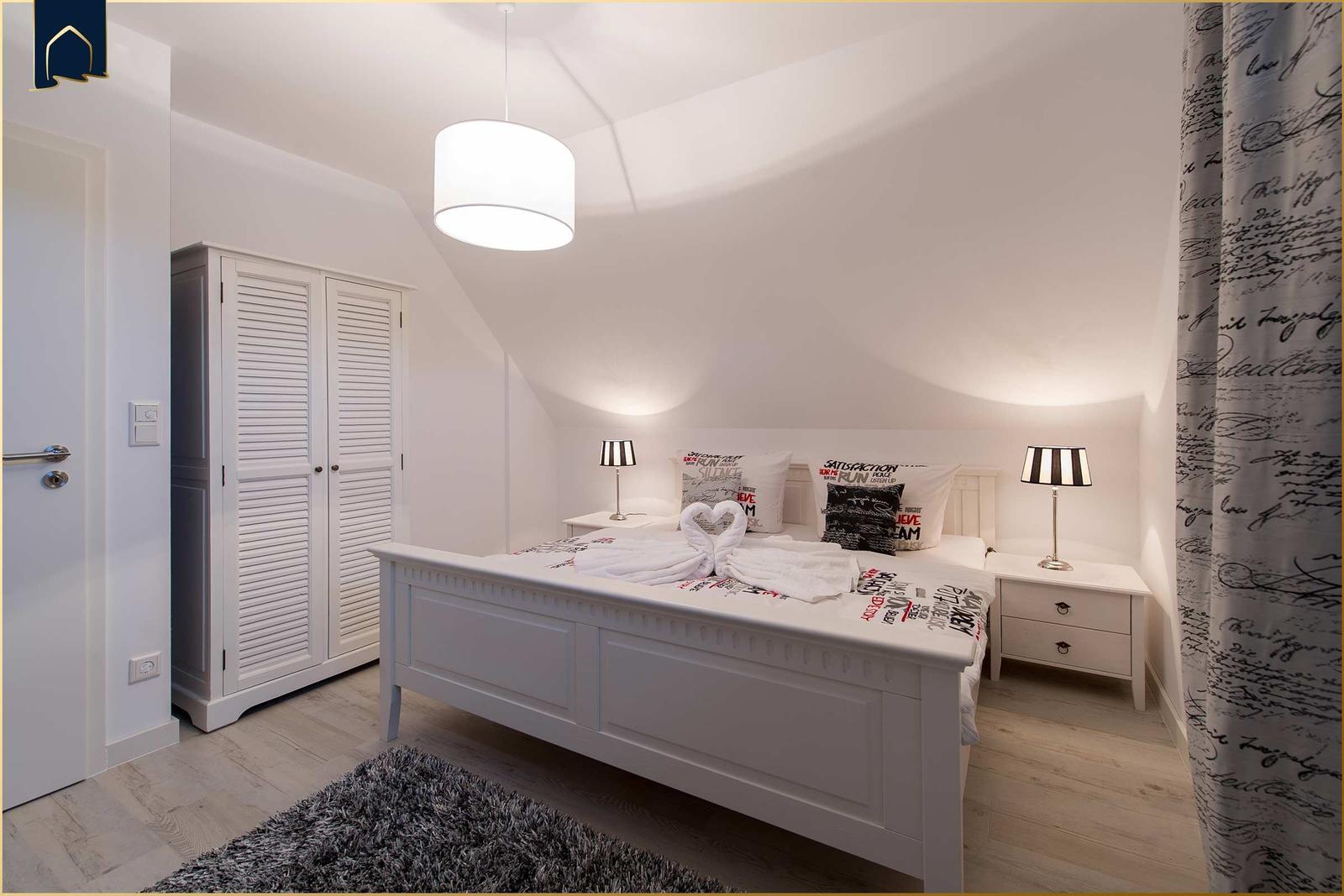 Schlafzimmer mit weißem Bett, Nachttischen und Kleiderschrank unter schrägem Dach.