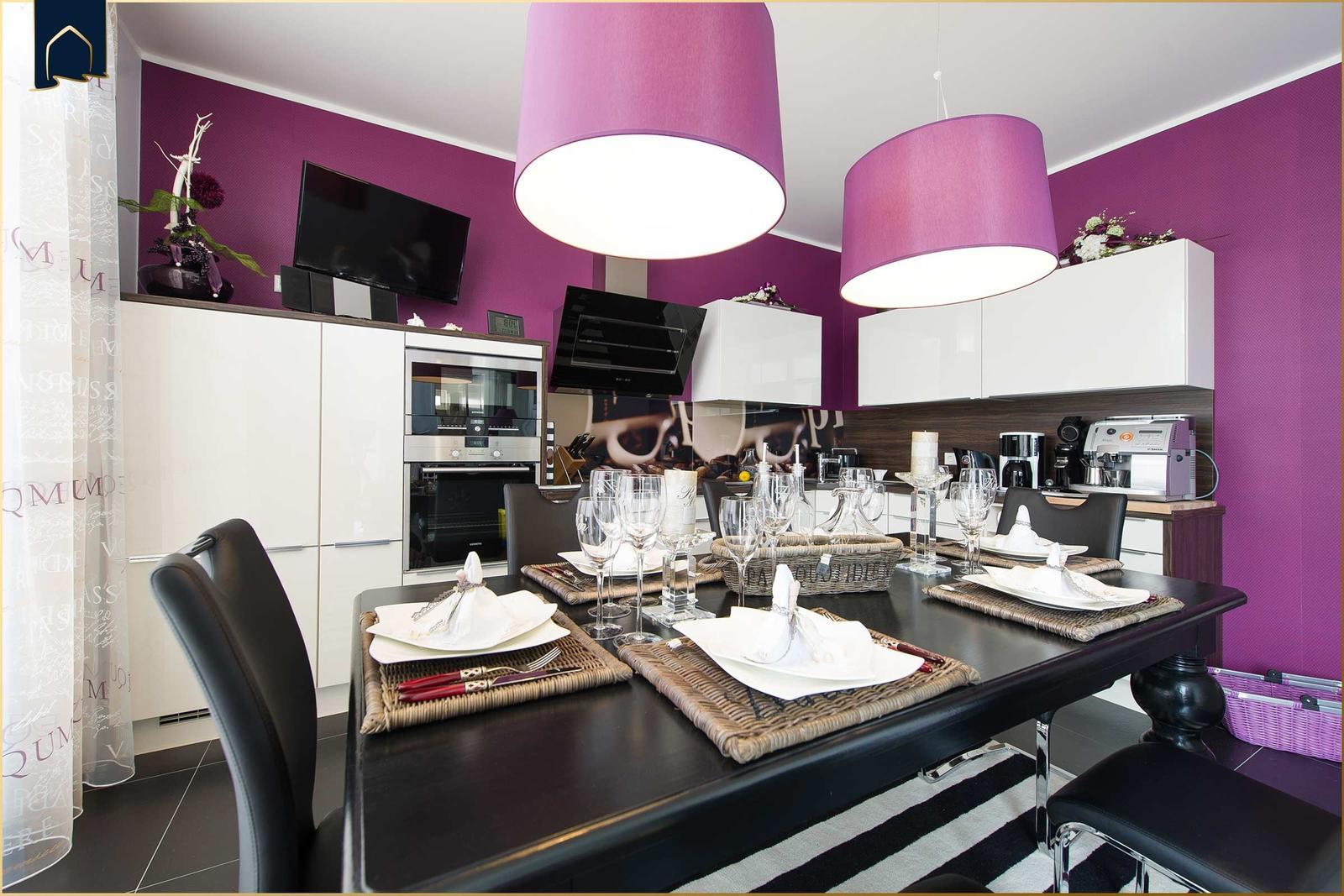 Essbereich mit Tisch, Stühlen und moderner Küche. Violette Wände und hängende Lampen.