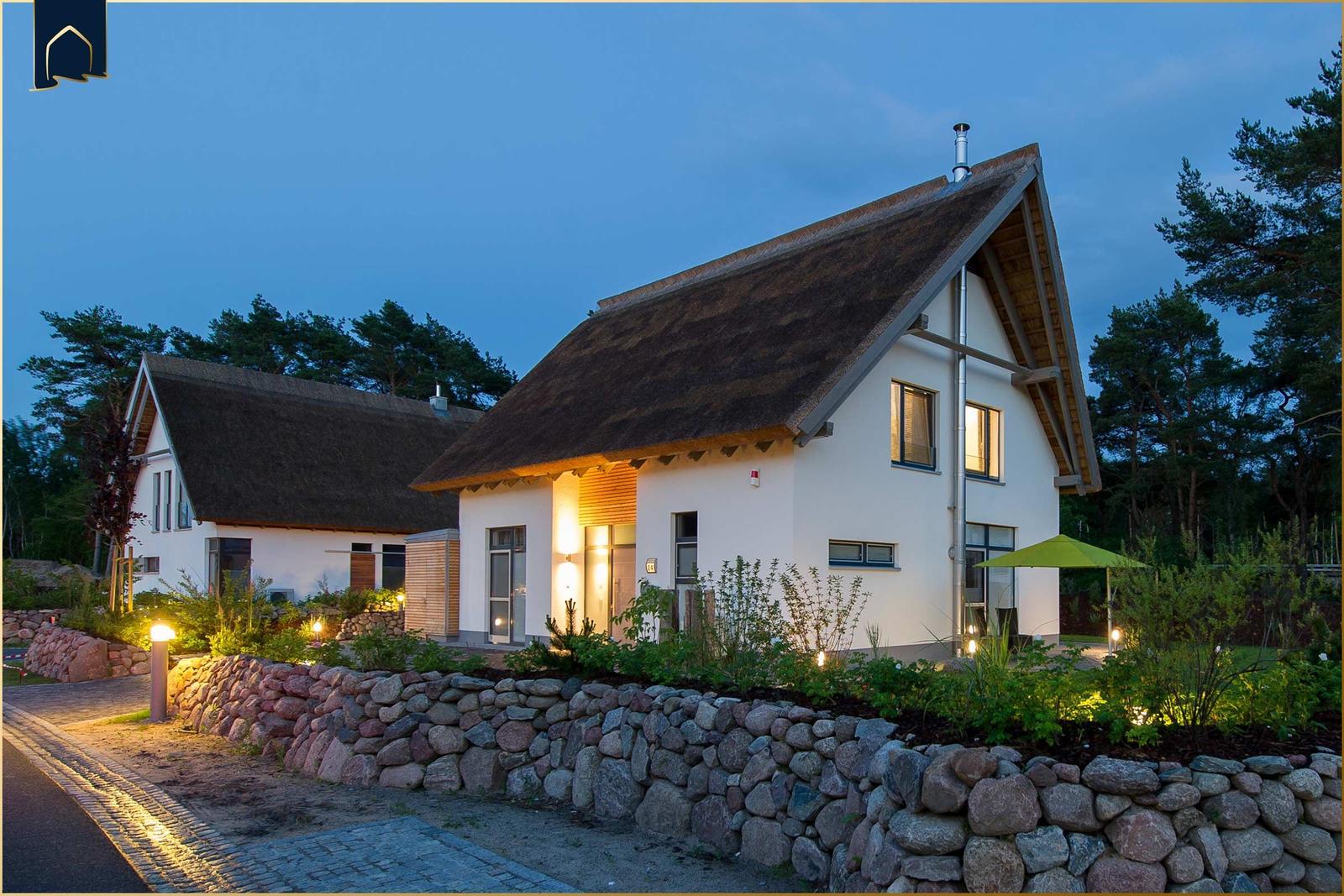 Haus mit strohgedecktem Dach, umgeben von Steinmauer und Bäumen bei Dämmerung.