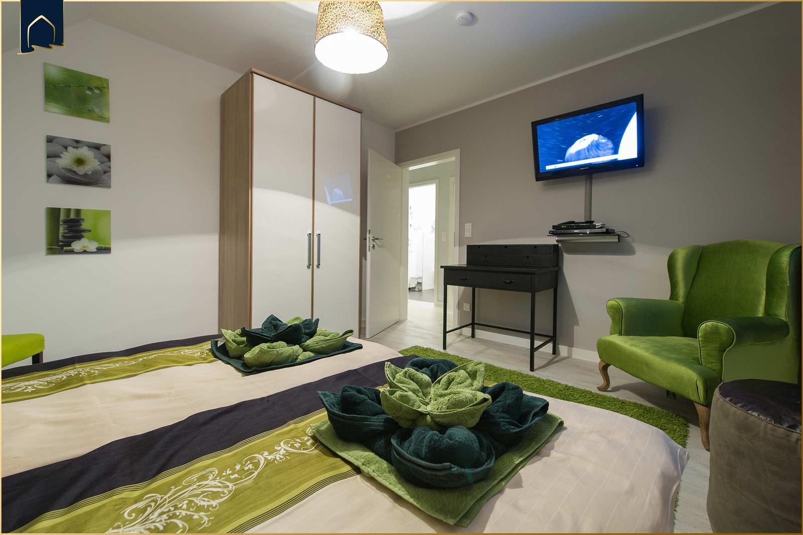 Schlafzimmer mit Bett, grüner Sessel und Wand-TV.