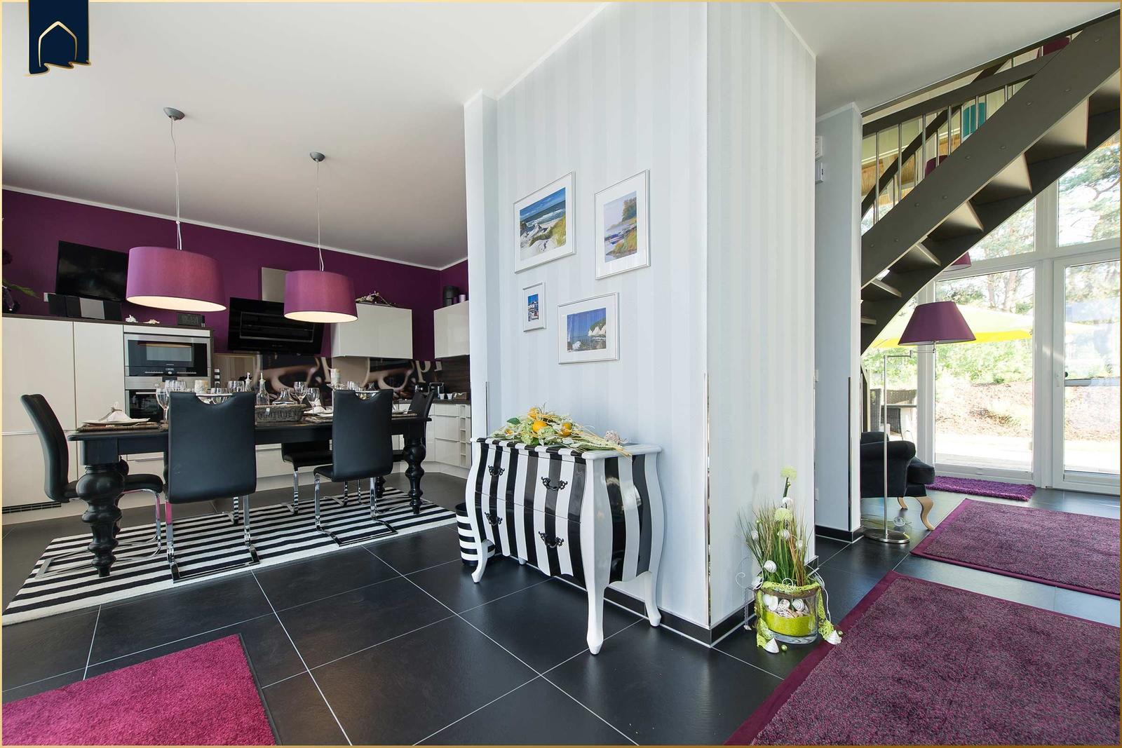 Essbereich mit Tisch, Stühlen und Küchenzeile. Treppenhaus und Wohnbereich sind sichtbar.