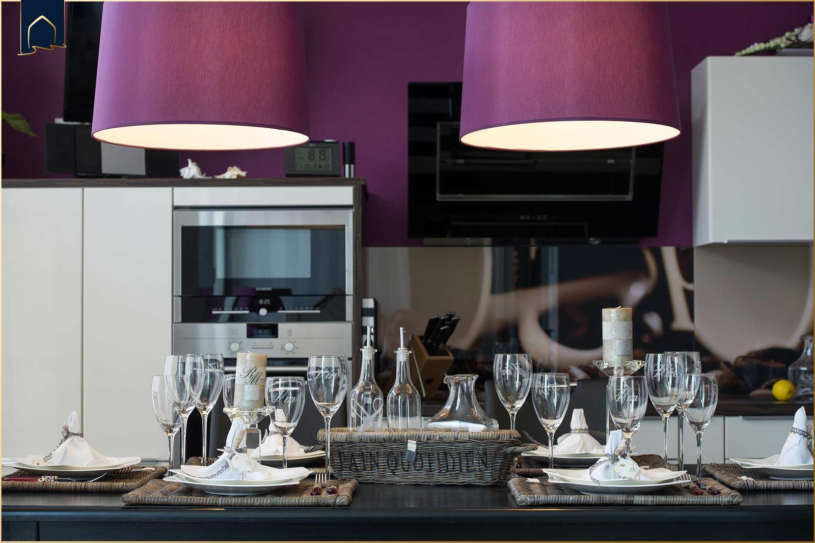 Gedeckter Esstisch mit Gläsern und Kerzen in moderner Küche mit lila Deckenleuchten.