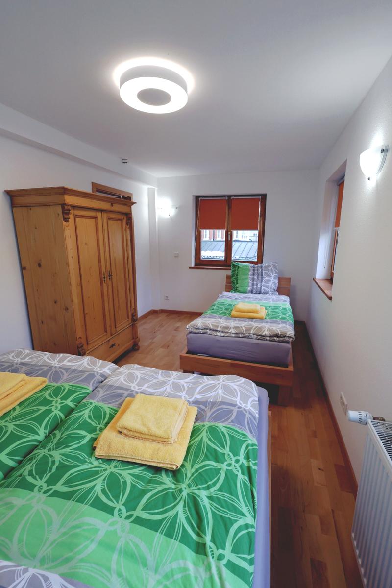 Zwei Betten mit grünem Bettzeug, Holzschrank und Fenster mit rotem Rollladen.