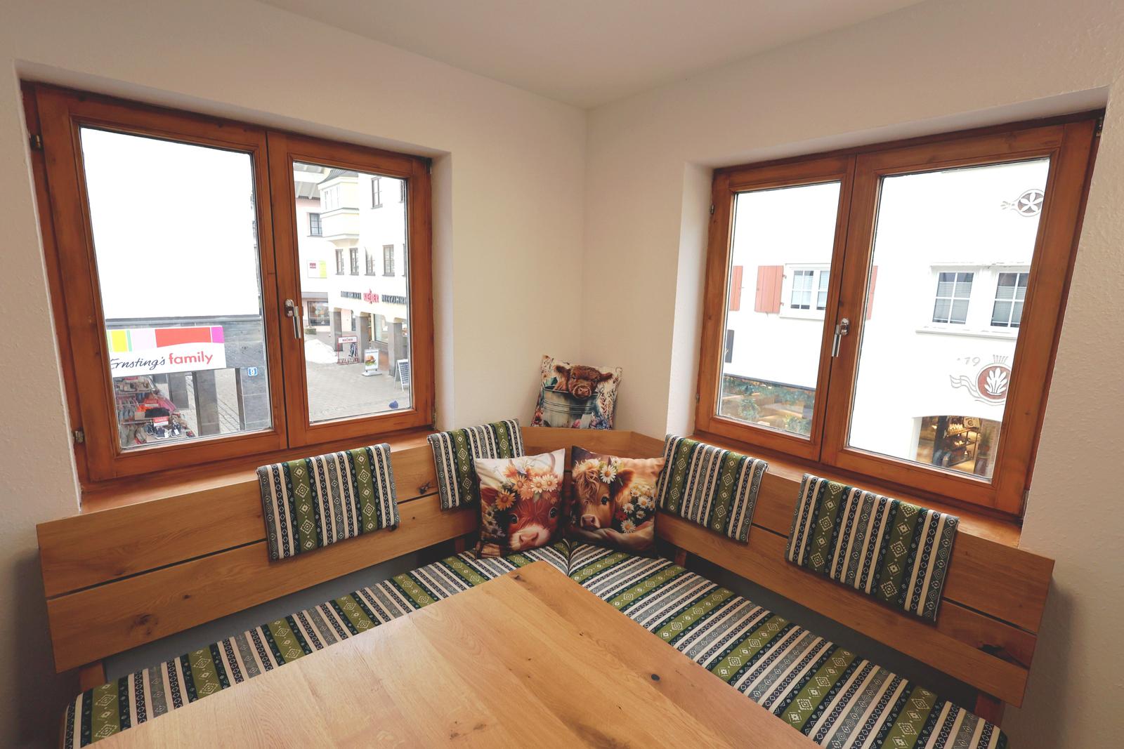 Eckbank mit Tischtuch und Kissen an zwei Fenstern.