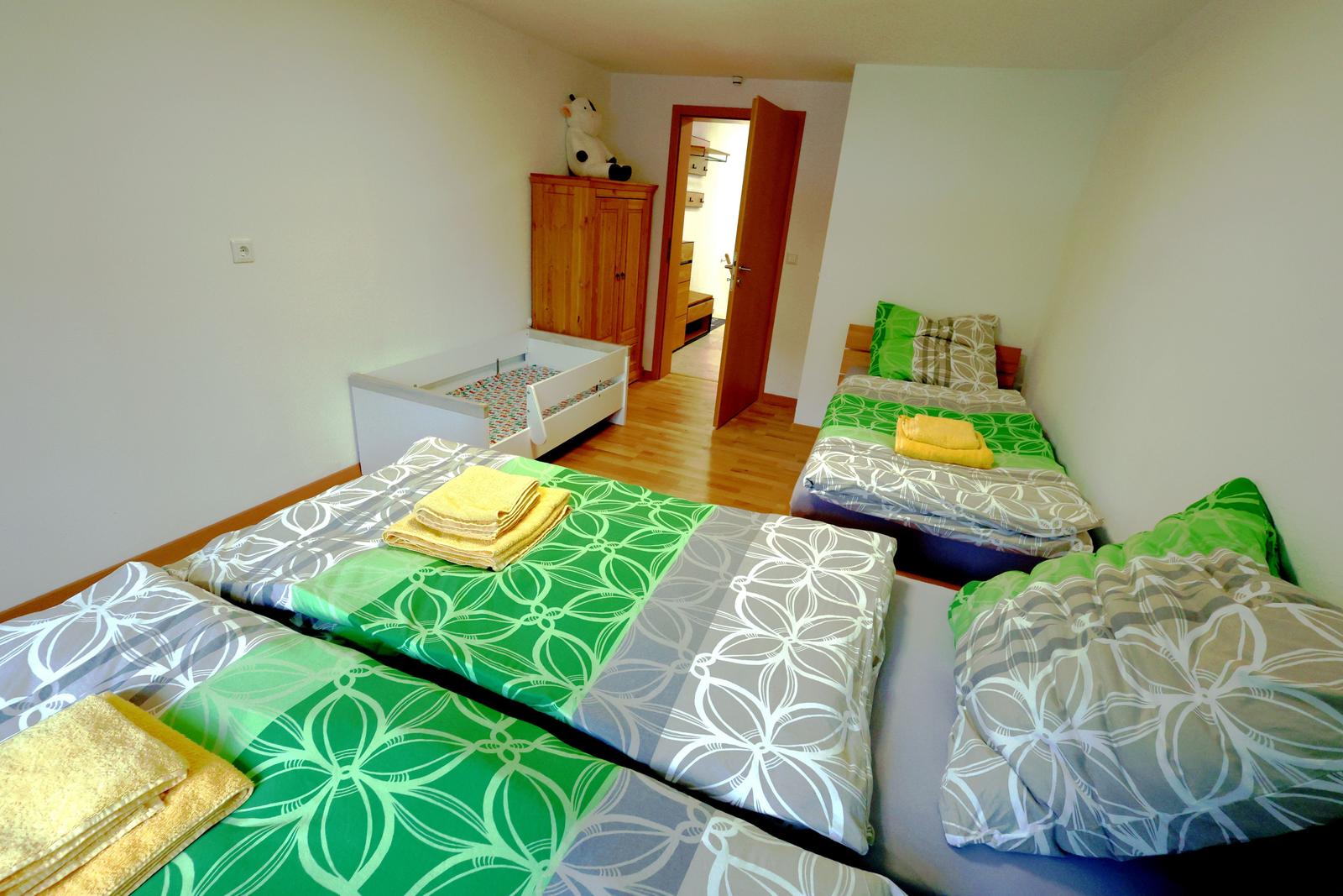 Zwei Betten mit grün-weißem Bettzeug und gelben Handtüchern. Ein Kinderbett und ein Schrank sind sichtbar.