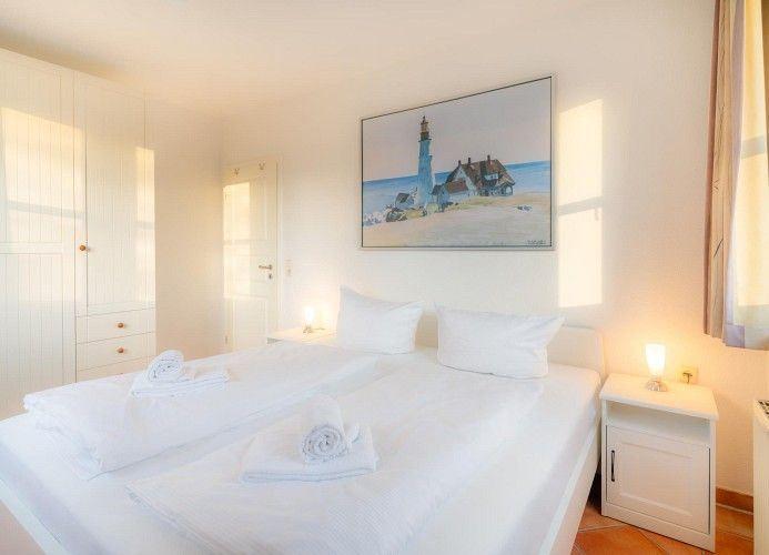 Schlafzimmer mit weißem Bett, Nachttisch und Wandbild mit Leuchtturm.