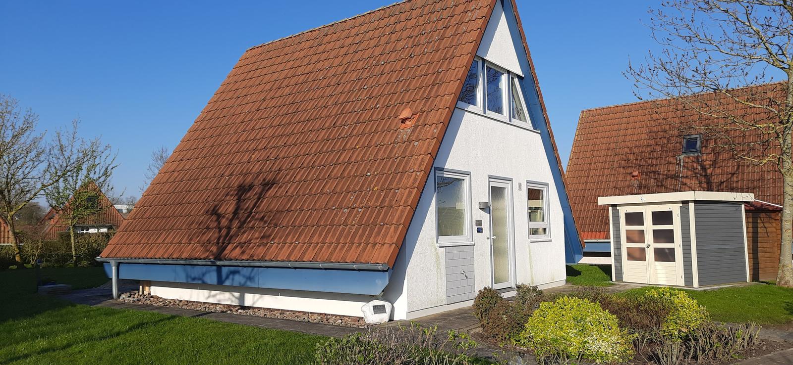 Ein Haus mit rotem Dach und weißer Fassade. Garten und kleiner Anbau sind sichtbar.