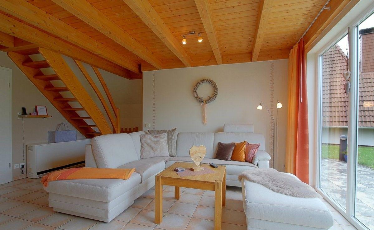 Wohnzimmer mit weißem Sofa, Holztisch und Holzbalkendecke. Große Fensterfront mit orangefarbenen Vorhängen.